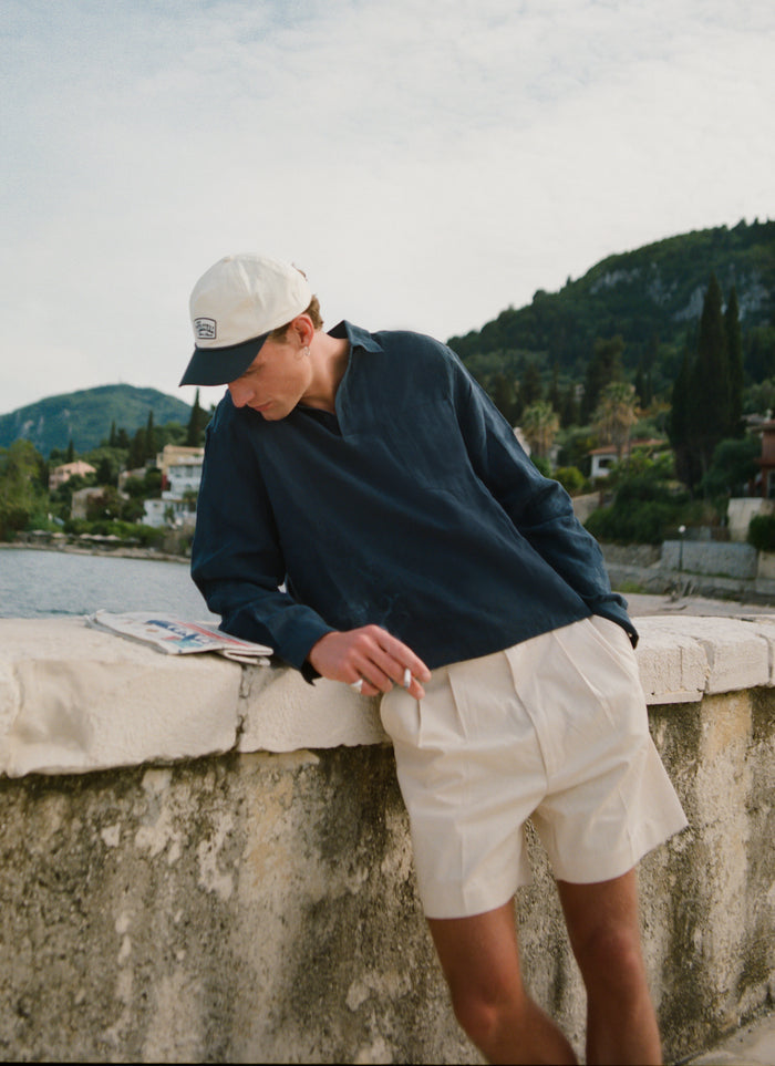 Navy Percival Alec linen pullover shirt in a boxy fit, worn by a model leaning against a stone wall by the sea.