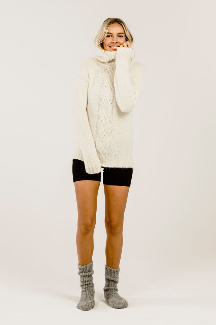 Woman wearing a cream cable knit sweater over a black pants with gray socks on a white background