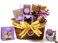 A low, oval wicker basket is tied with a yellow congratulations ribbon. The basket holds one home assortment chocolate box, butter crunch in a purple window box, one boxed chocolate bar, a bag of chocolate espresso beans, a thank you bar in a gold box with a brown ribbon, salted caramels in a purple window box, a 7” chocolate champagne bottle.