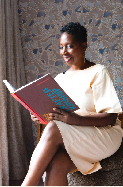 Woman reading a book titled 'Crafted Kinship' against a patterned wall.