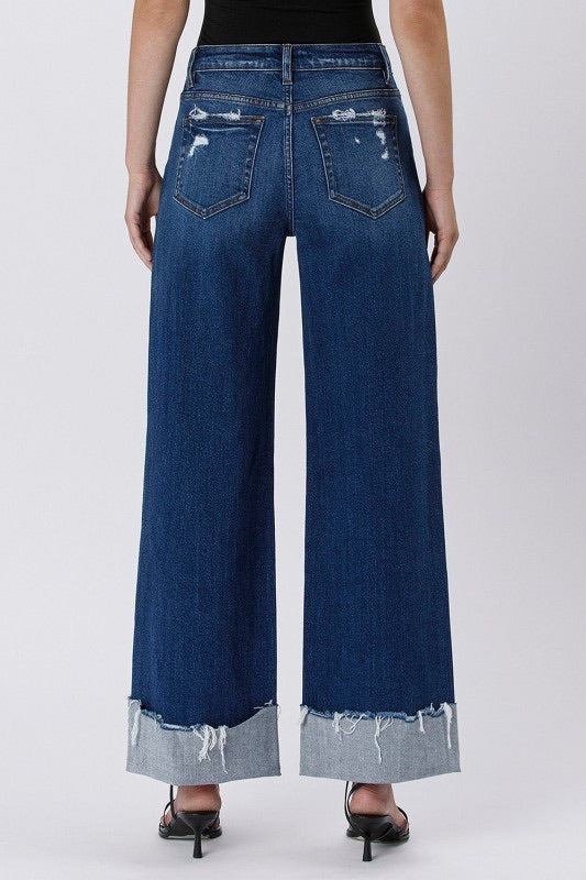 Model wearing high rise cuffed baggy wide leg jeans with slight distressing and raw single cuff hem, styled with black heels and a fitted black tank top (back view).