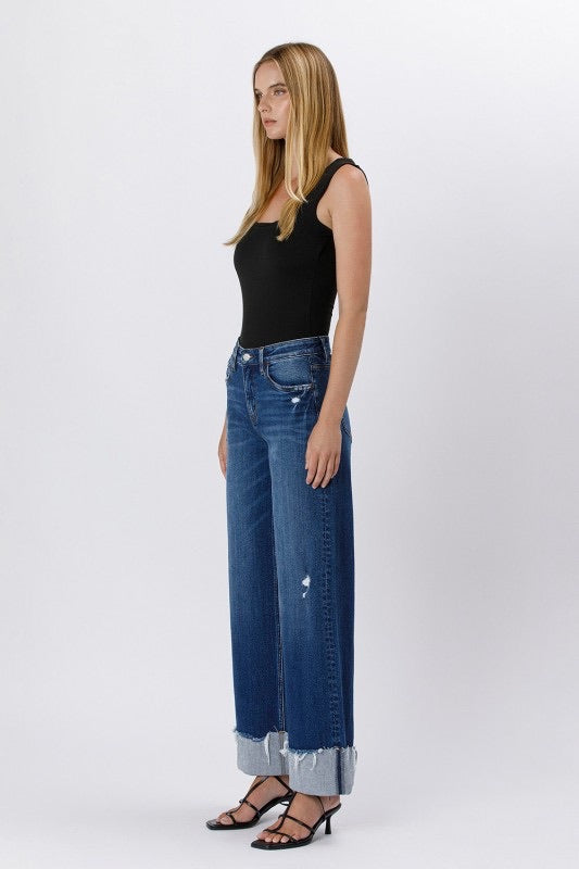 Model wearing high rise cuffed baggy wide leg jeans with slight distressing and raw single cuff hem, styled with black heels and a fitted black tank top (side view).