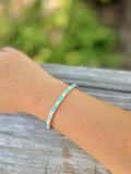 Gold bracelet with turquoise band and sparkle accents on a wrist with blurred background.
