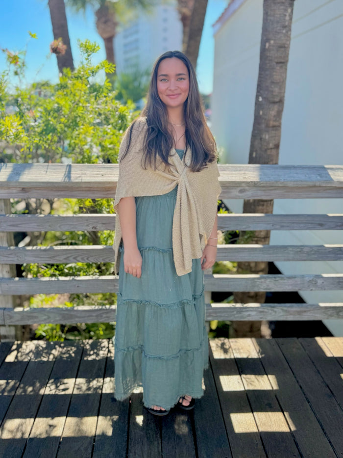 Woman in a green dress with a gold shawl standing on a wooden deck with trees and a building in the background.