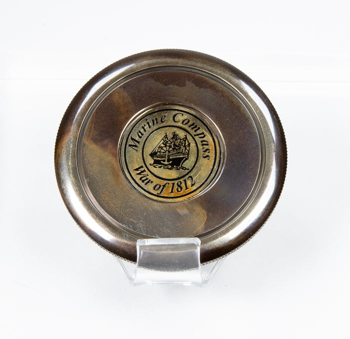 Close up shot of compass cover with engraved image of ship encircled by the words: "Marine Compass War of 1812"