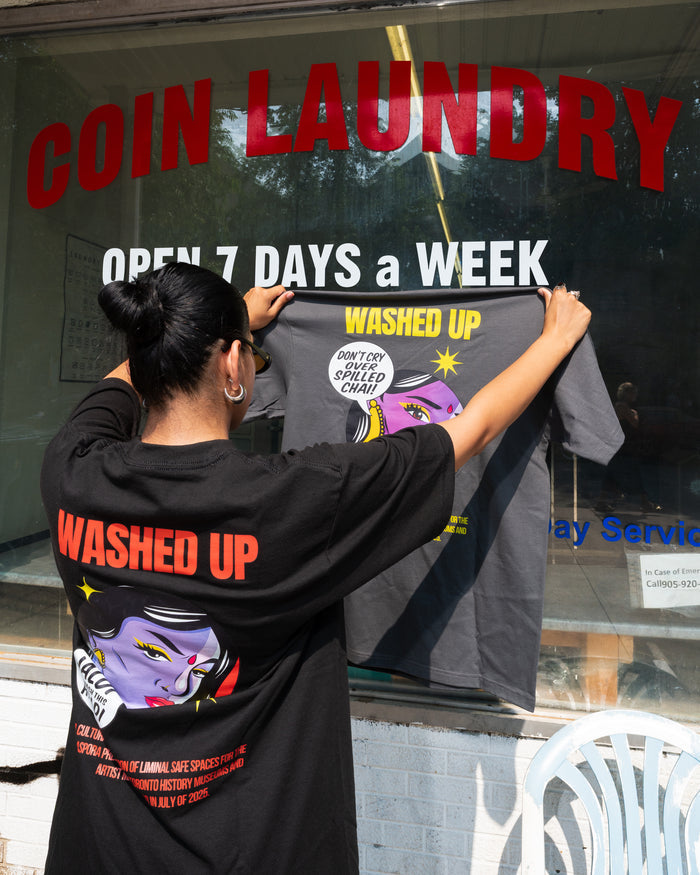 Person holding a t-shirt with 'WASHED UP' branding in front of a coin laundry sign.
